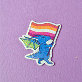 A sticker that has a cute dragon carrying a lesbian pride flag. This pride sticker makes a great gift
