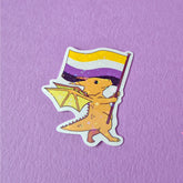A sticker that has a cute dragon carrying a non-binary / enby pride flag. This pride sticker makes a great gift