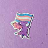 A sticker that has a cute dragon carrying a trans pride flag. This pride sticker makes a great gift