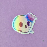Holographic skull sticker with a stealth trans coronation flower on it. Makes a great pride gift