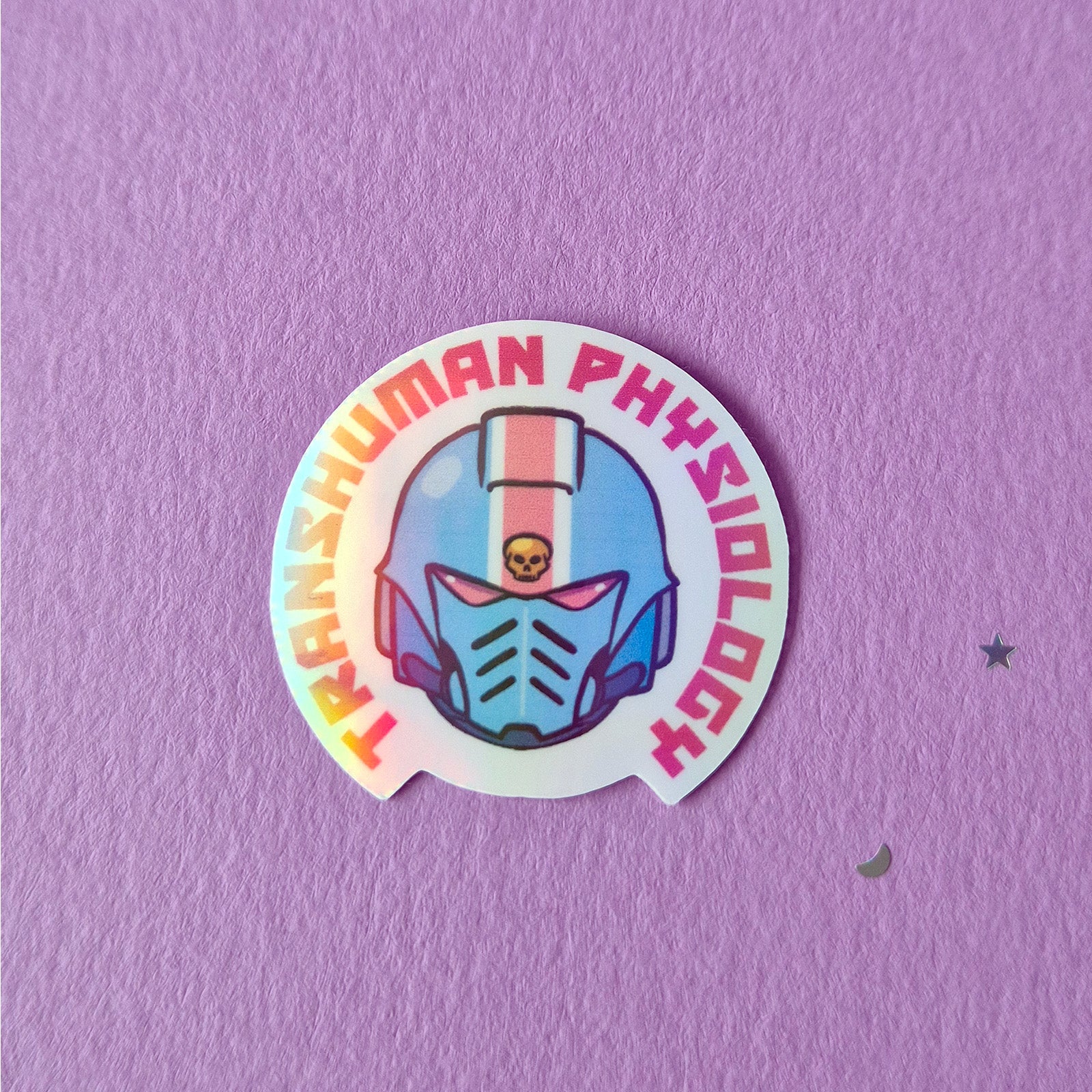 A holographic finish sticker that reads 'Transhuman physiology'. Featuring a 40k helmet in trans colours.