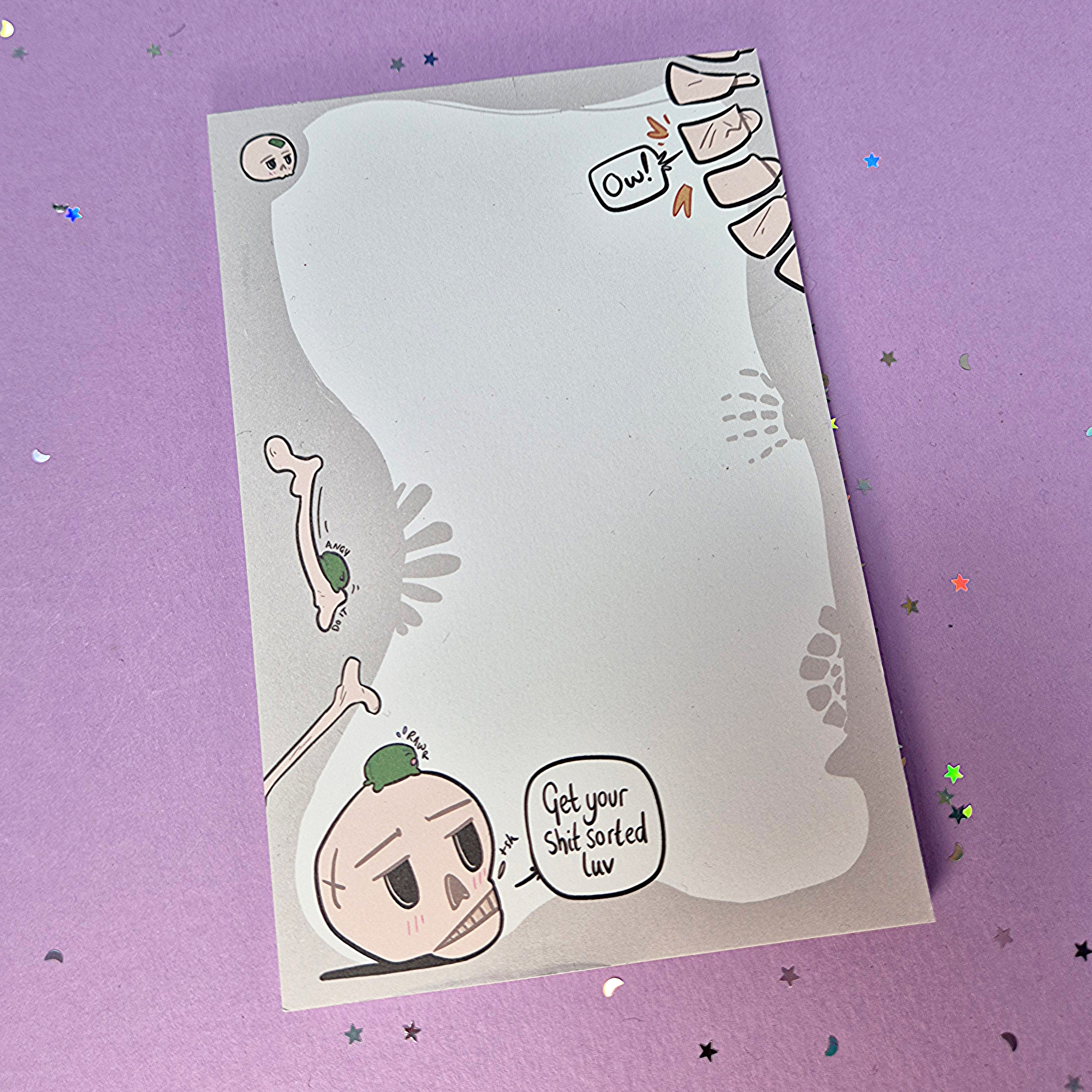 Memo pad with decorative border showing various skeleton and skulls. Top right has a spine that is cracking with 'Ow!' speech bubble coming from it. Bottom left has a skull with a small frog on it. Skull has speech buble saying 'Get your shit sorted luv,'