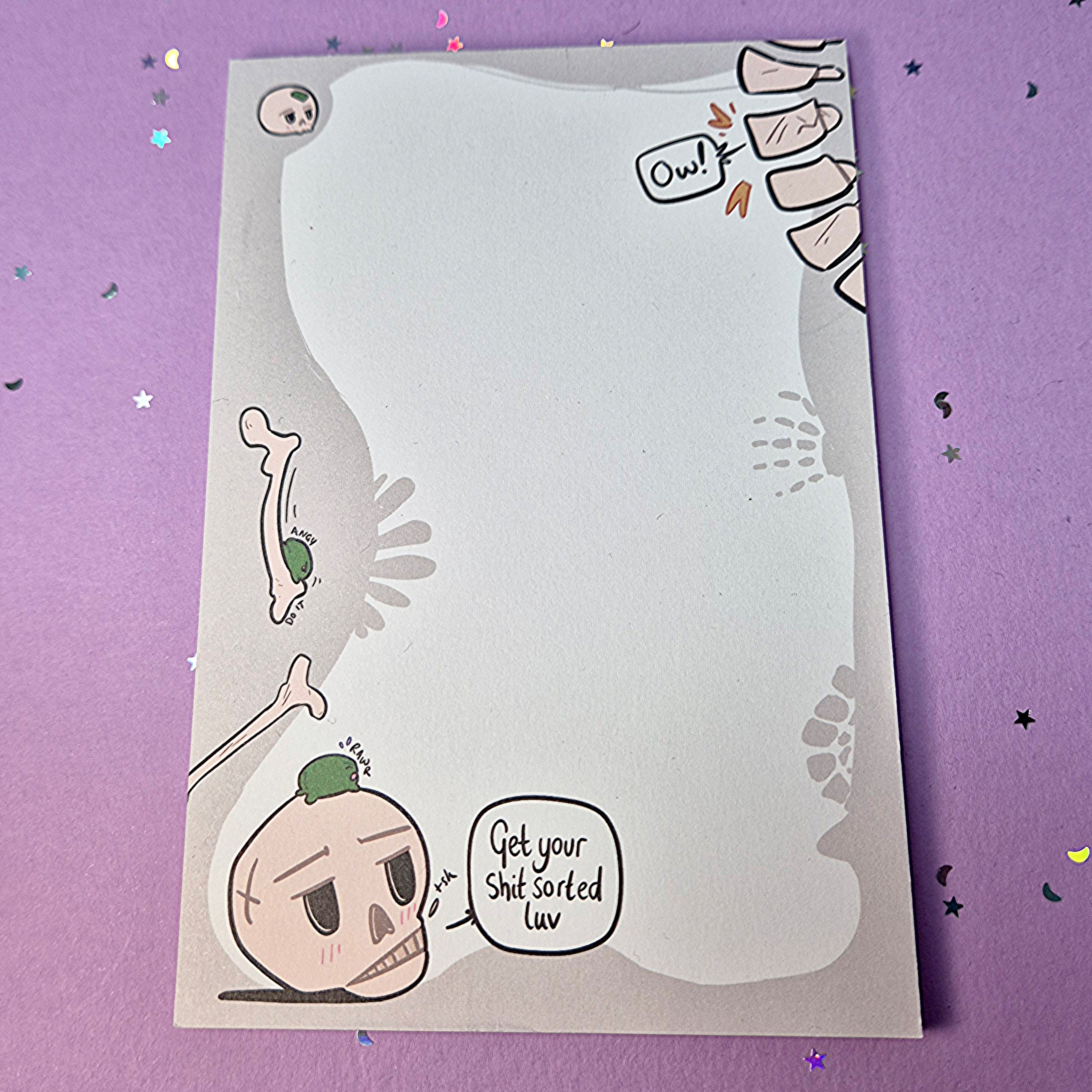 Memo pad with decorative border showing various skeleton and skulls. Top right has a spine that is cracking with 'Ow!' speech bubble coming from it. Bottom left has a skull with a small frog on it. Skull has speech buble saying 'Get your shit sorted luv,'